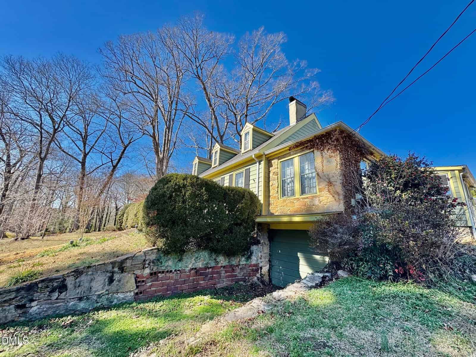 A two-story yellow house with dormer windows and green trim sits on a grassy lot with bare trees, overgrown bushes, and a stone retaining wall under a clear blue sky.