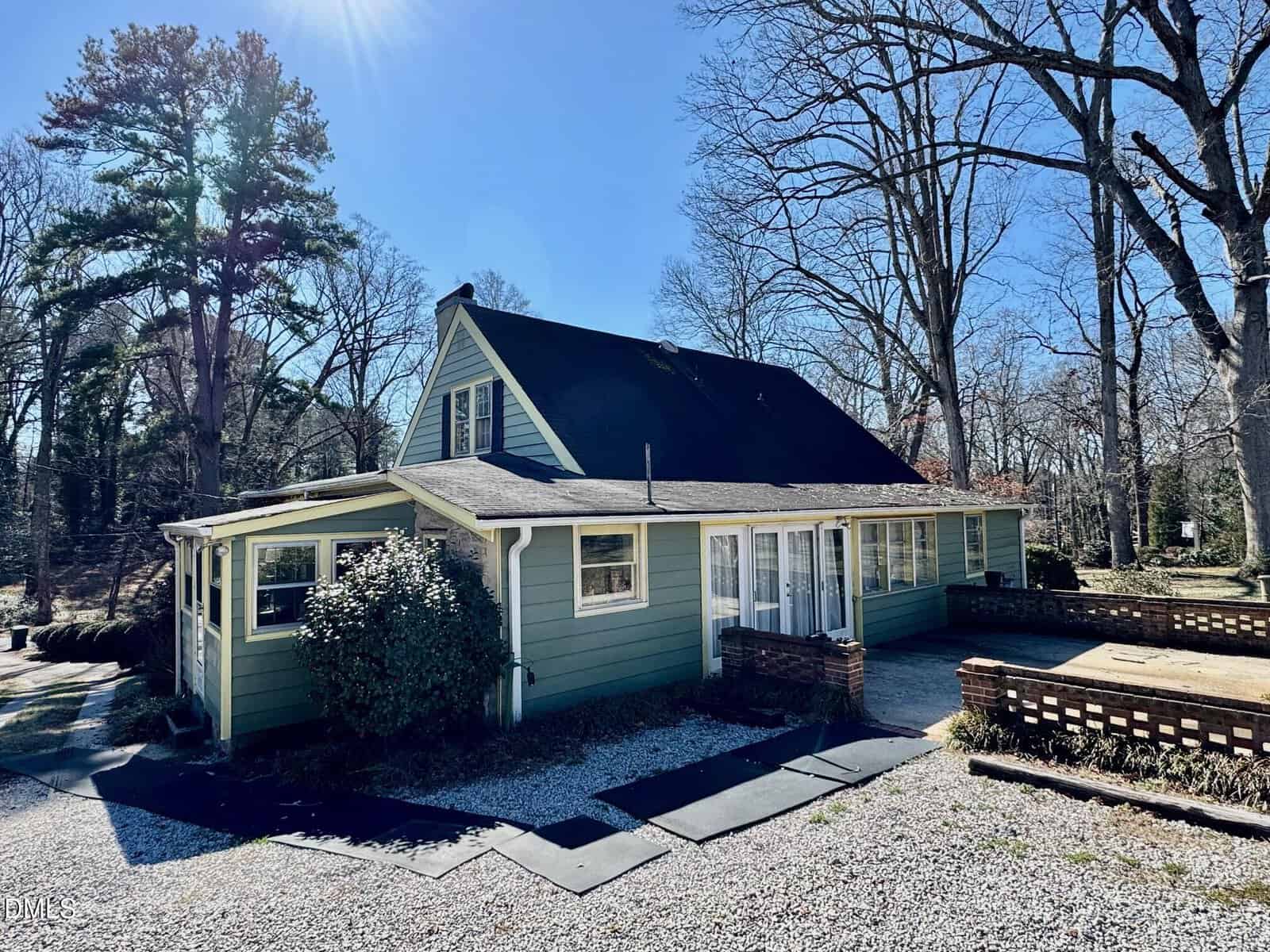 A green two-story house with a steep, dark roof sits in a wooded area. Sunlight shines from the top left. A gravel driveway and small brick patio are in front, with leafless trees and shrubs surrounding the property.