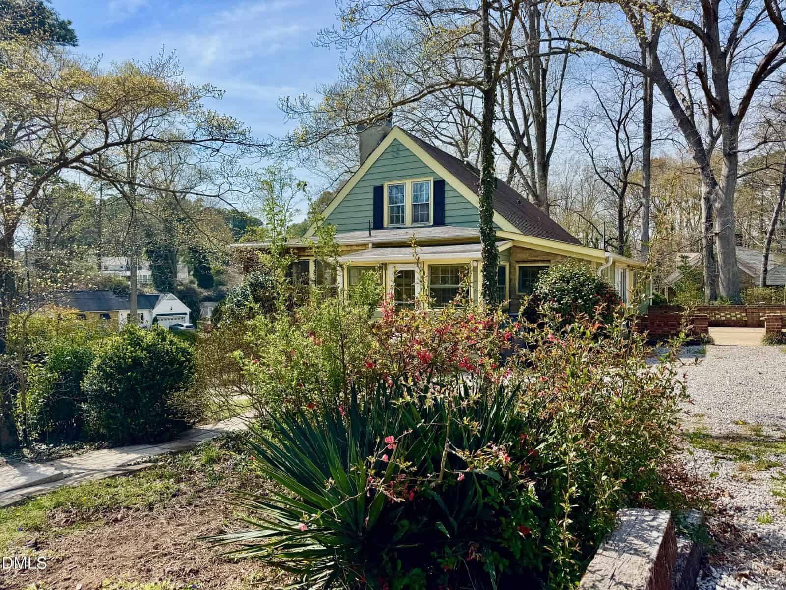 A small green-and-yellow house with a covered front porch sits among tall trees and dense shrubs on a sunny day, with a gravel driveway to the right and a garden in the foreground.