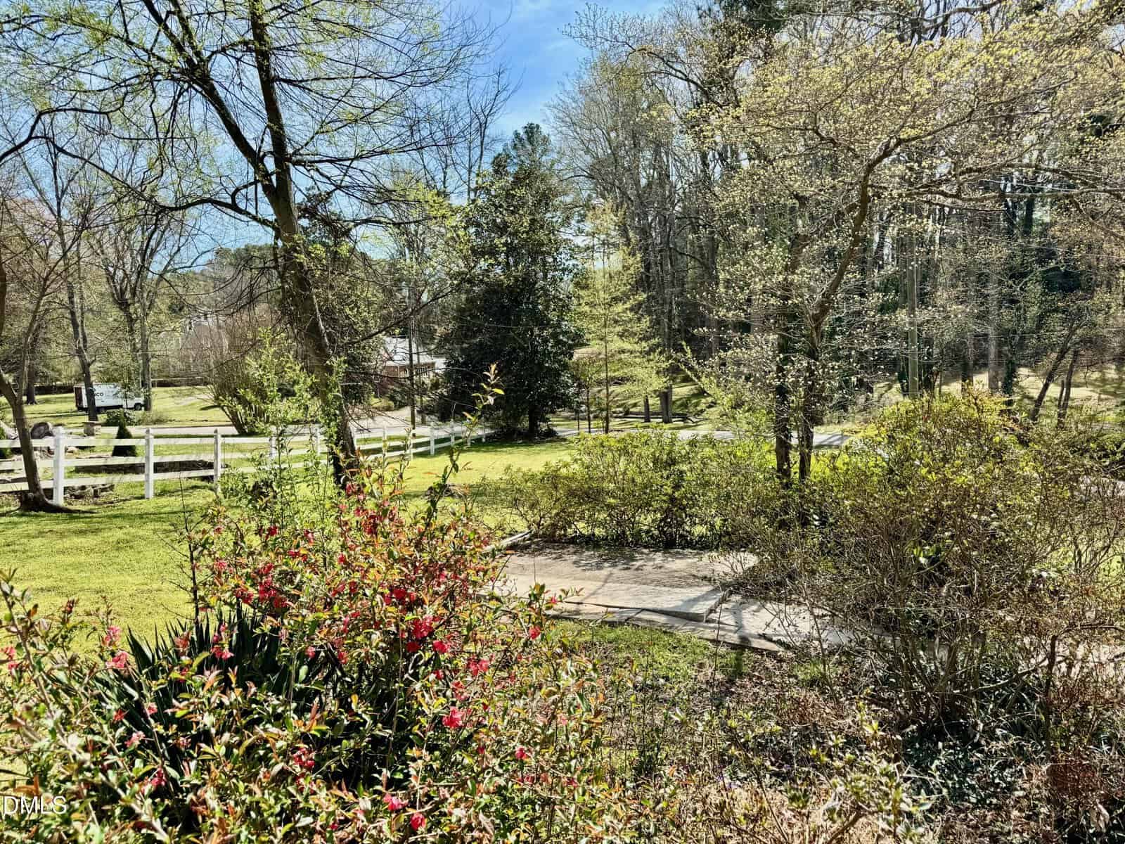 A sunny yard with blooming pink flowers, green shrubs, and trees. A white wooden fence borders the grassy area. A paved path curves through the scene. Houses and tall trees are visible in the background under a blue sky.