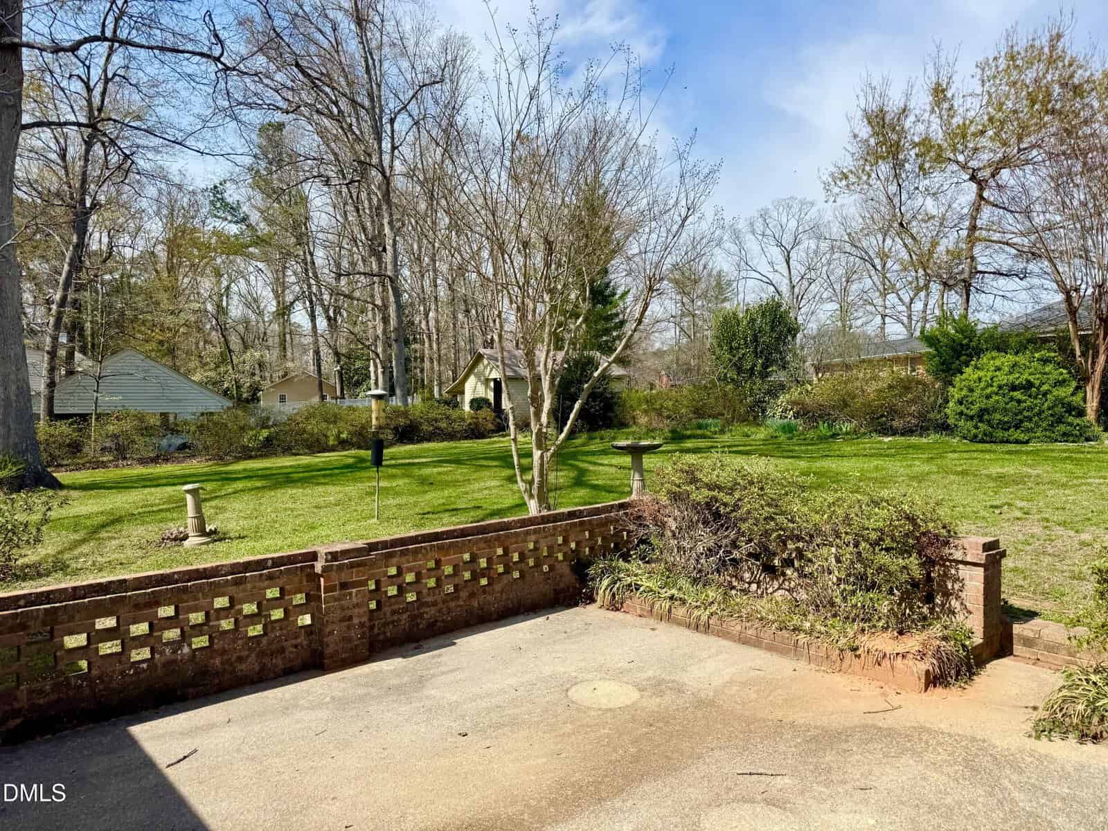 A backyard with a grassy lawn, mature trees, shrubs, and a small brick patio area. Houses and a garden shed are visible in the background. A birdbath and a bird feeder stand in the lawn. The sky is clear and sunny.