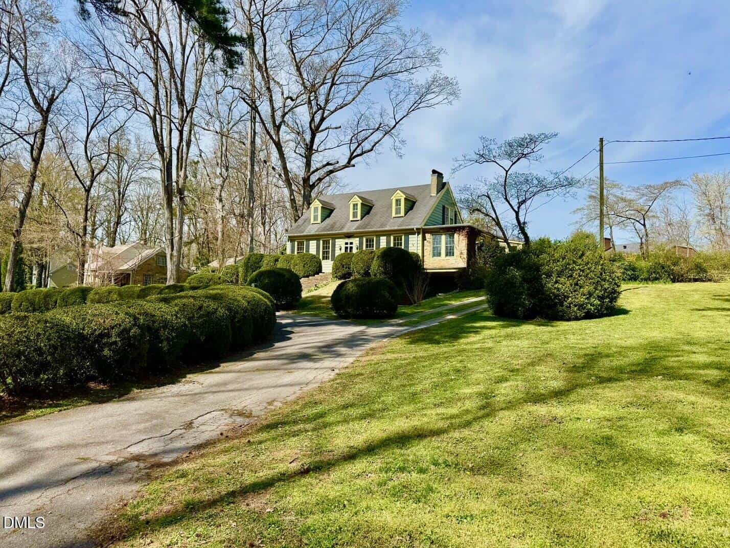 A large house with a dark roof, yellow dormer windows, and stone accents sits on a grassy lawn surrounded by trees and neatly trimmed bushes on a sunny day. A driveway curves up toward the entrance.