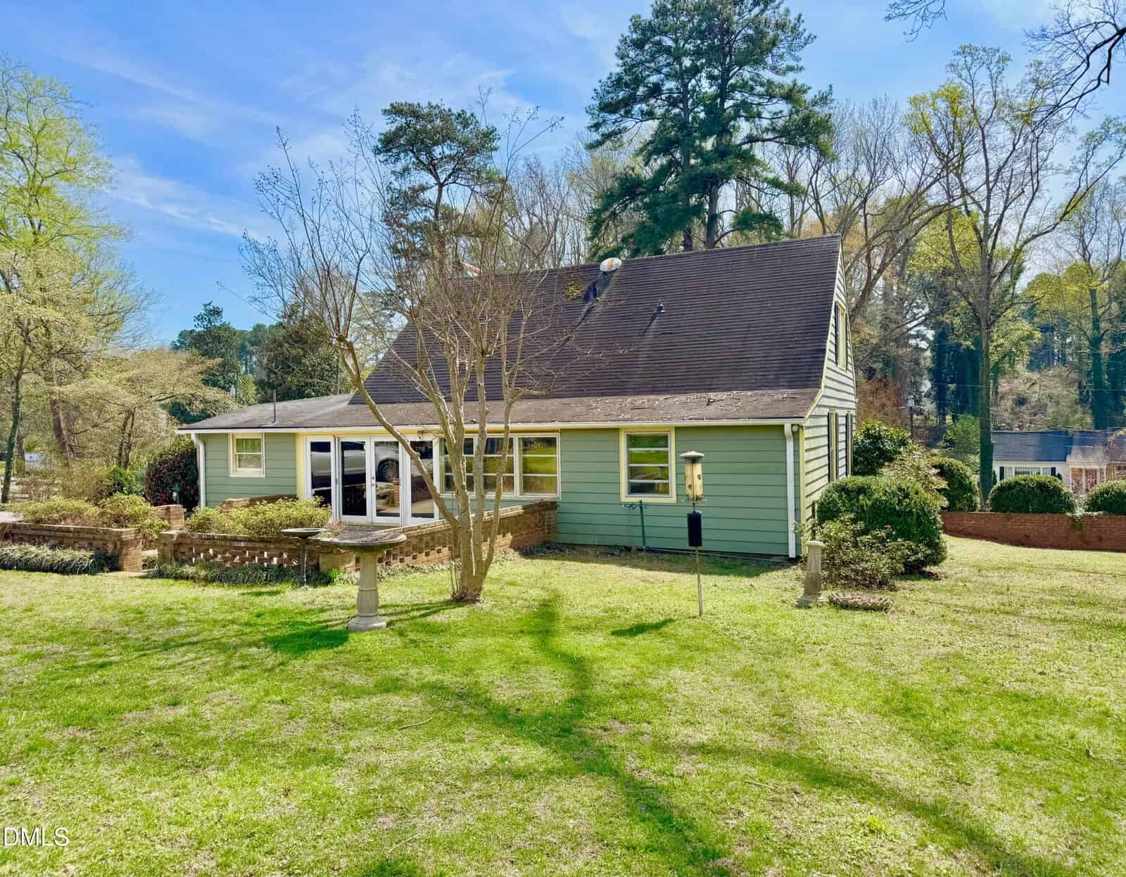 A light green house with a dark roof sits on a grassy lawn, surrounded by trees and bushes. A leafless tree, birdbath, and bird feeder are in the foreground under a clear, blue sky.