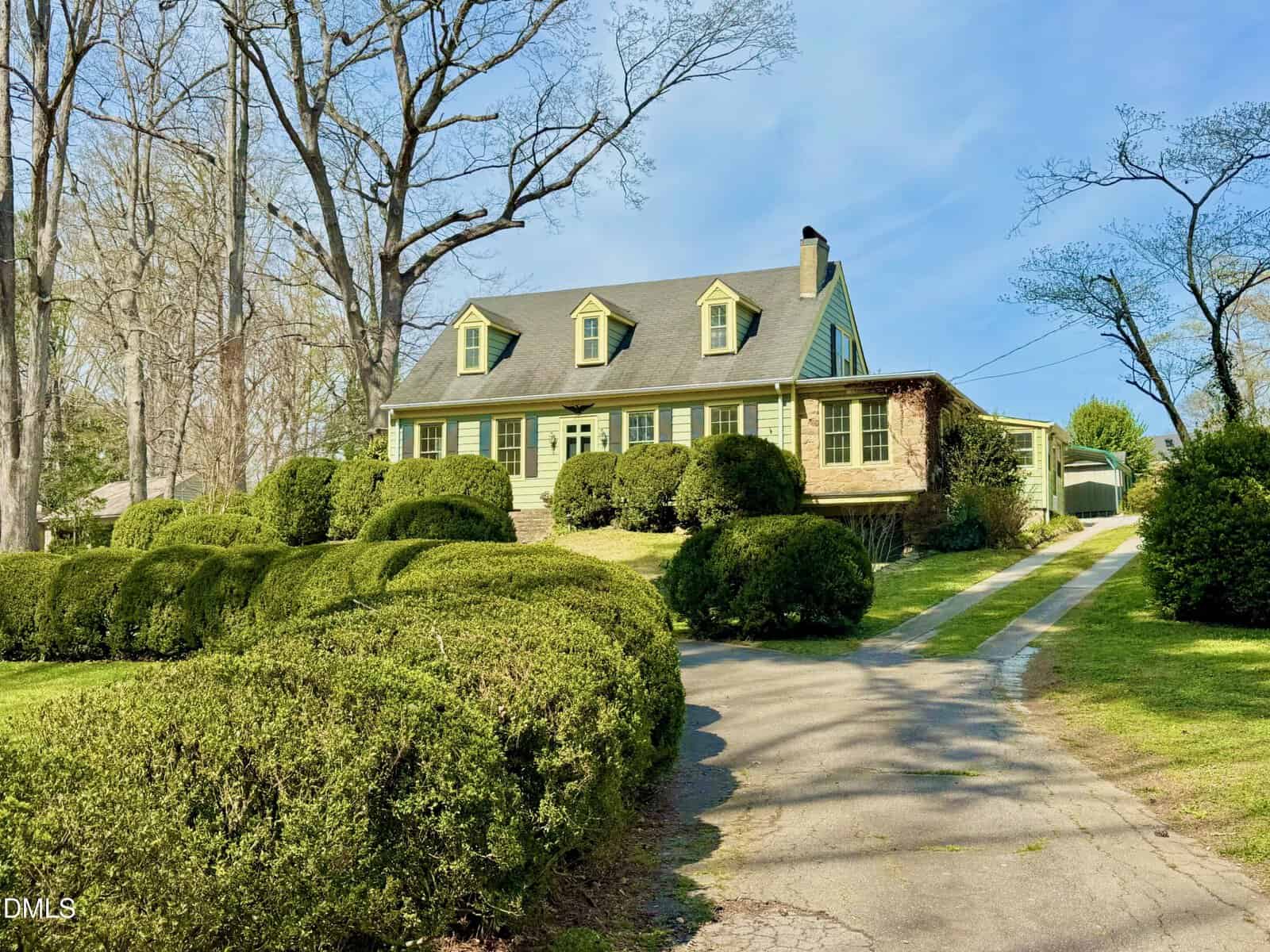 A two-story house with green shutters and dormer windows sits on a grassy hill at 222 Willowood Drive, Henderson, surrounded by trimmed bushes and tall leafless trees. A driveway leads up to the house, with a detached garage visible in the background.