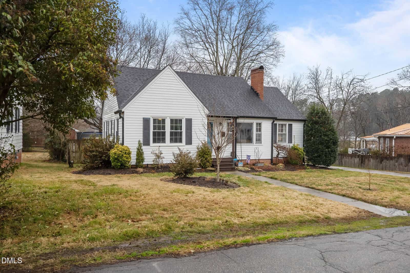 One-story white house with black shutters and a dark roof, featuring a brick chimney and small front porch. The yard has grass, shrubs, and trees, with a concrete walkway leading to the porch.