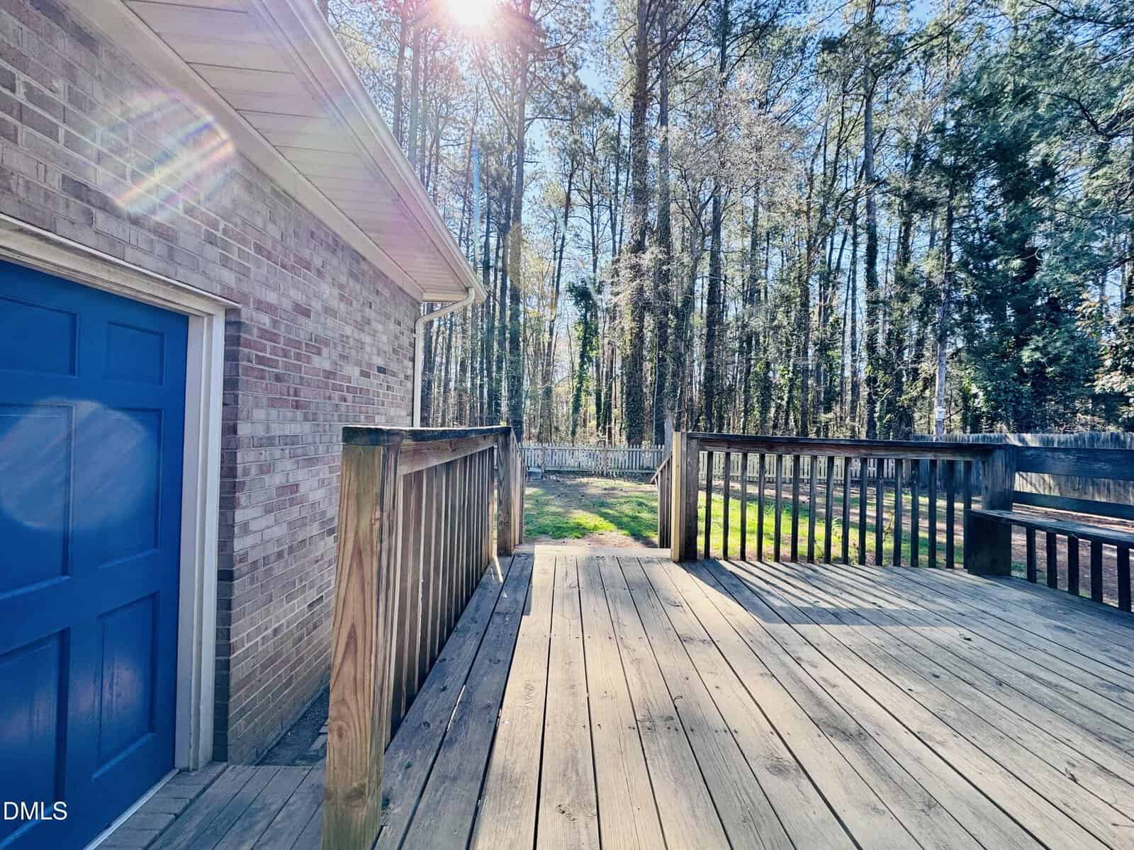 A backyard deck with wooden railings attached to a brick house with a blue door, overlooking a grassy yard and tall trees under bright sunlight.