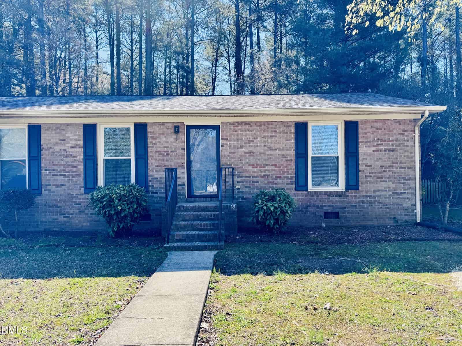 Charming single-story brick house located at 1205 W C Street, Butner, featuring black shutters, a central front door with a glass storm door, two bushes flanking the entrance, and a concrete walkway leading up to four brick steps. Trees are visible in the background.