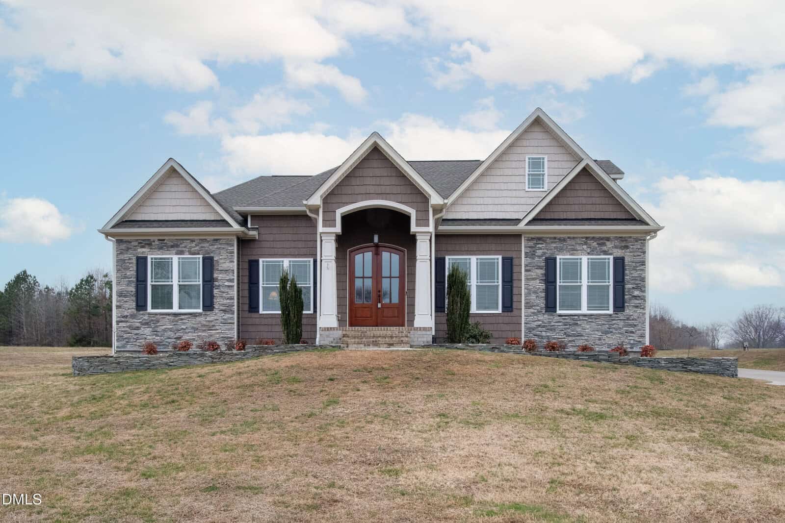 Single-story house with gray stone and brown siding, white trim, and a symmetrical façade. There are double wooden front doors, two front windows with shutters, and small bushes by the entrance. The sky is partly cloudy.
