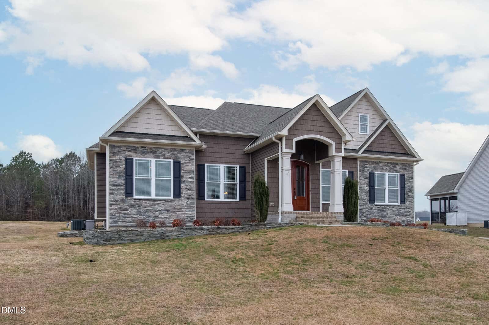 Single-story house with stone and siding exterior, three front-facing gables, white-trimmed windows, a wooden front door with an arched portico, and a lawn with sparse grass under a partly cloudy sky.