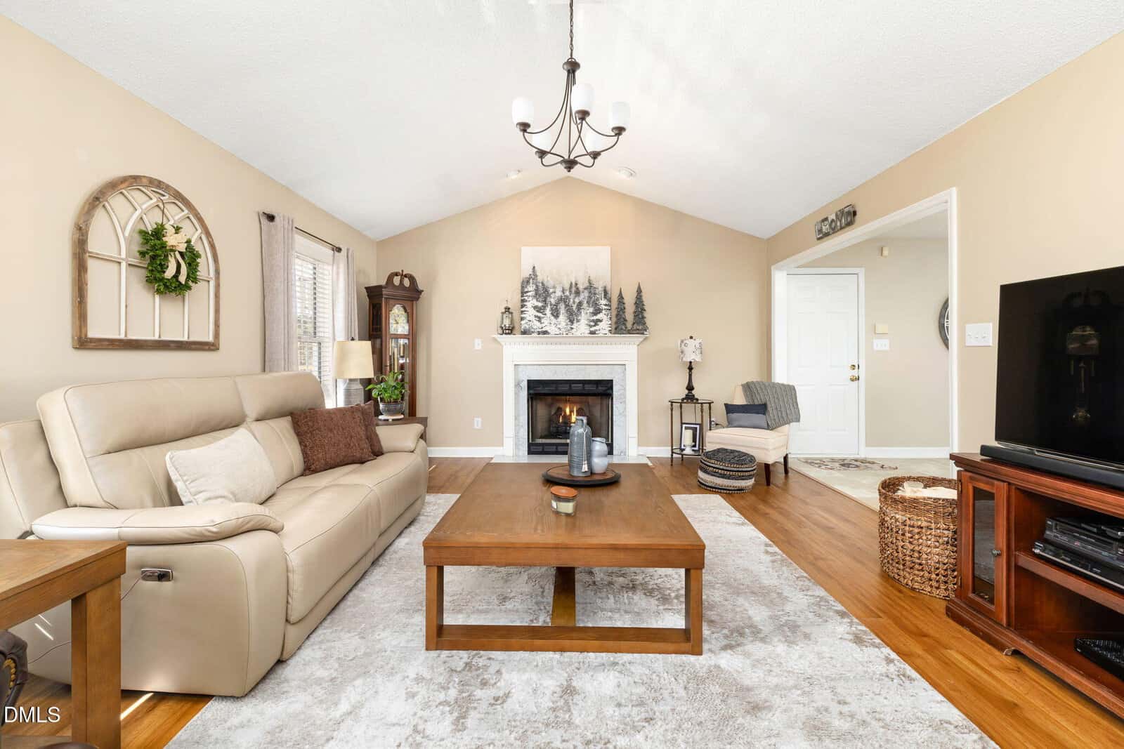 A living room with beige walls, a cream sofa, a wooden coffee table, a fireplace with decor above it, a TV on a stand, a window with curtains, and a light fixture on the ceiling.