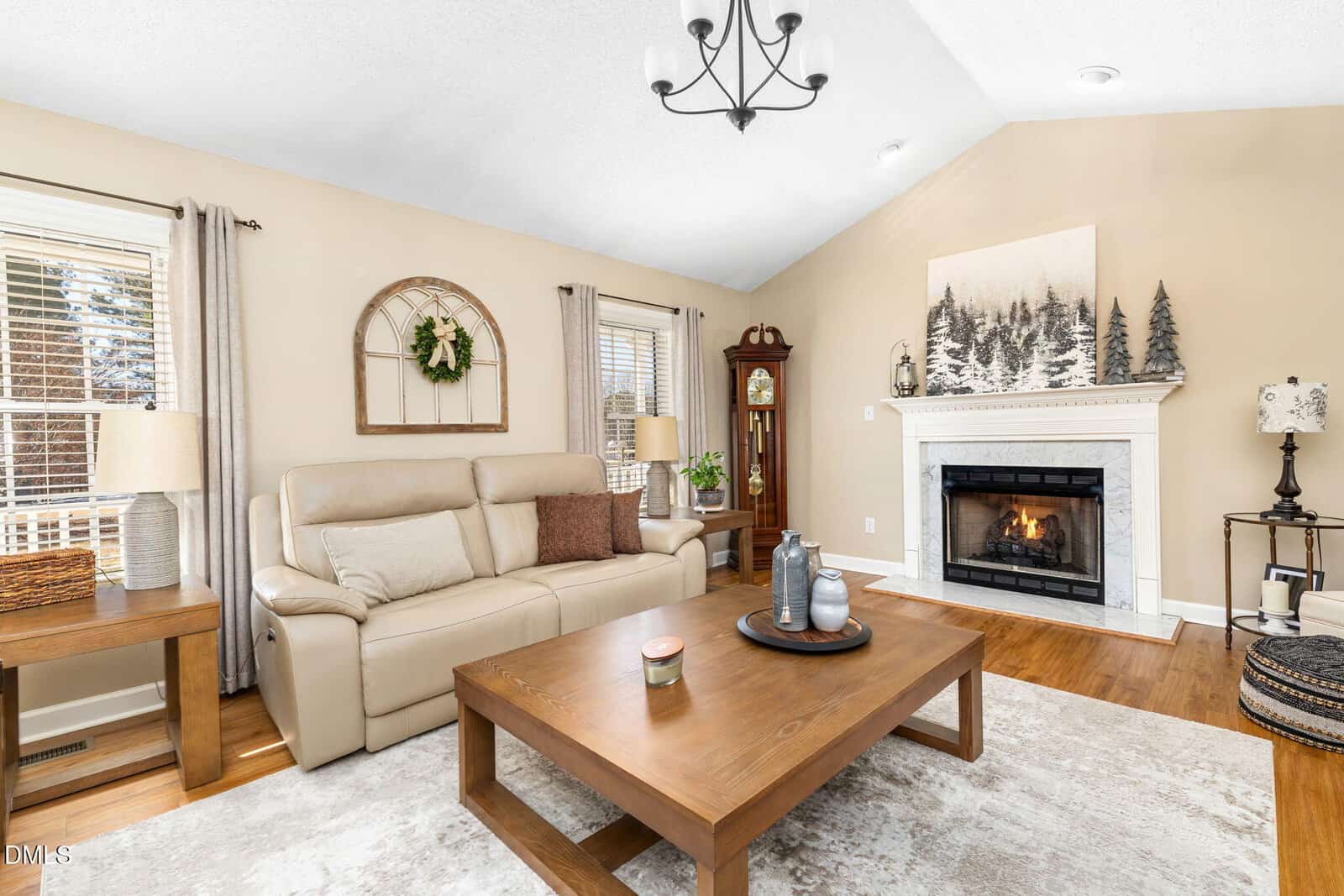 Living room with beige walls, a cream sofa, wooden coffee and side tables, a grandfather clock, a lit fireplace with art above it, and two windows with light curtains. Hardwood floors and a neutral rug complete the space.