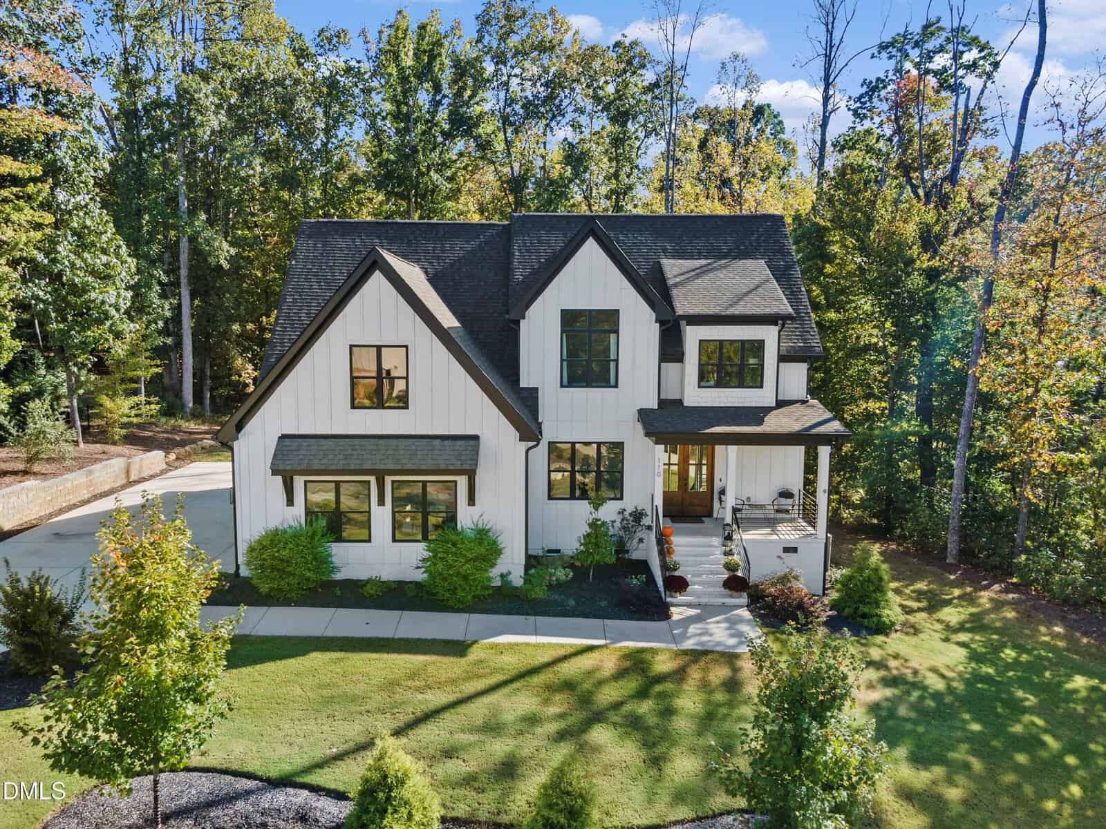 Two-story modern farmhouse with white vertical siding and black trim, surrounded by green grass, shrubs, and trees at 110 Black Swan Drive in Youngsville. Concrete driveway and front walkway lead to a covered porch with outdoor furniture. Blue sky and sunlight in the background.
