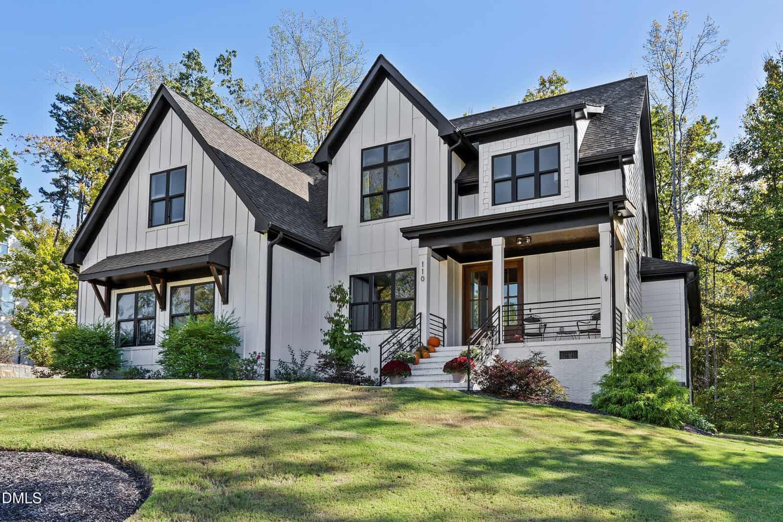 A modern two-story house with white vertical siding, black trim, large windows, and a covered front porch. The yard is grassy with a few shrubs and trees in the background under a clear blue sky.