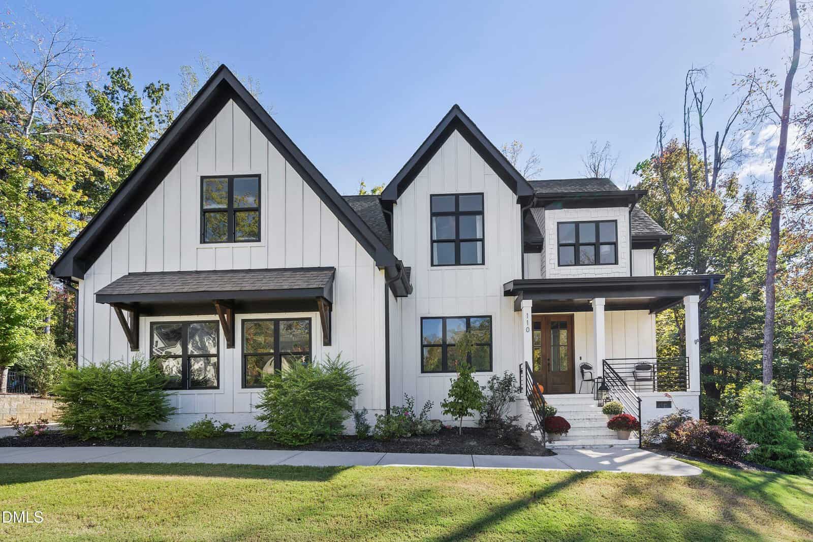 A modern two-story white house features black trim, steep gable roofs, a covered front porch with railing, and a landscaped yard with bushes and trees. The home is set against a backdrop of tall trees and a clear blue sky.