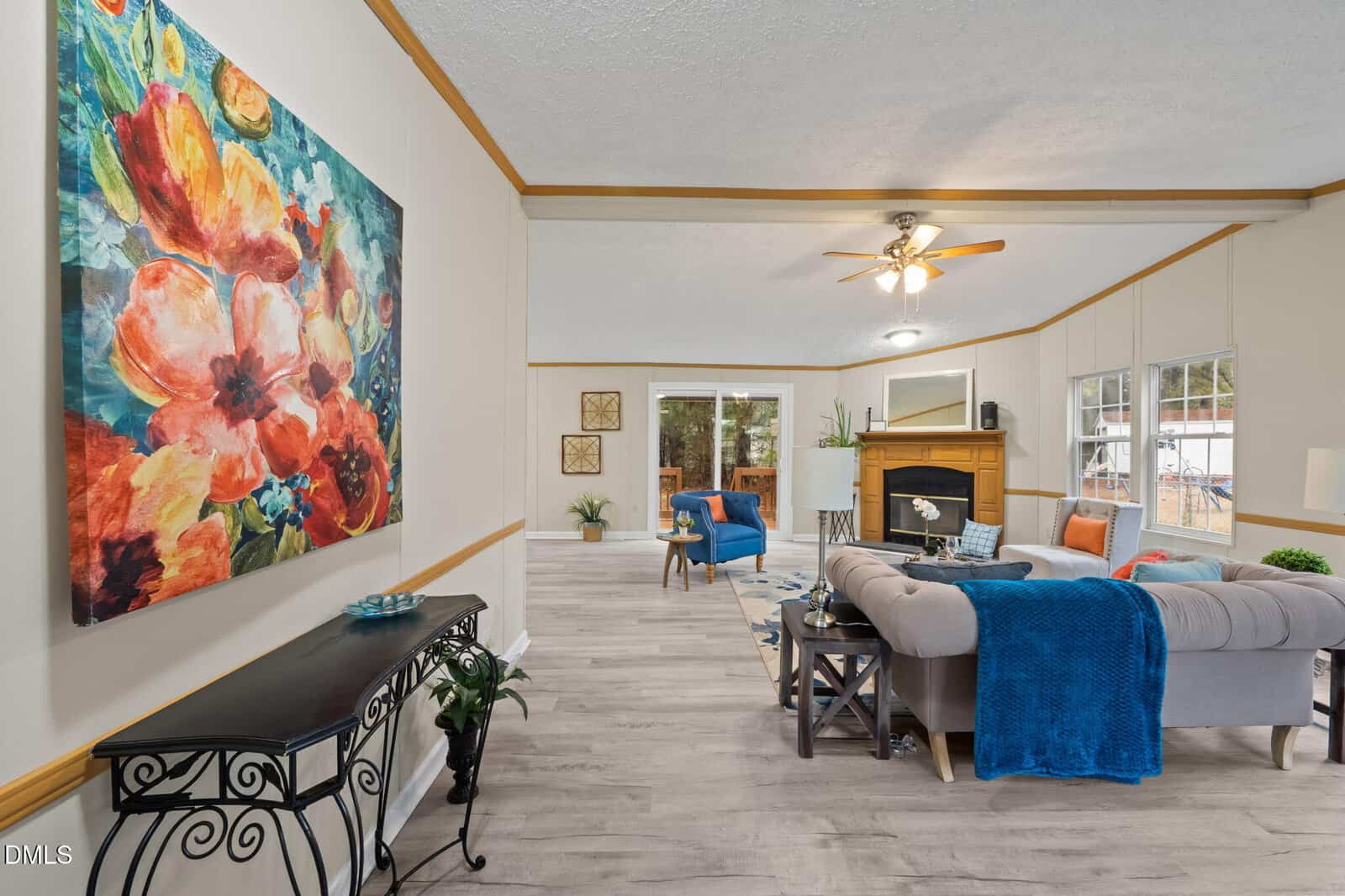 A living room with light-colored walls and floors, a ceiling fan, a beige sectional sofa with blue accents, a fireplace, large windows, and a colorful floral painting on the left wall.