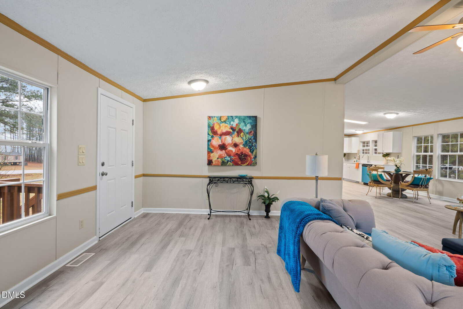 A living room with light wood flooring, a gray sofa with blue accents, a table lamp, a console table beneath a colorful flower painting, and an open view into a dining area and kitchen.