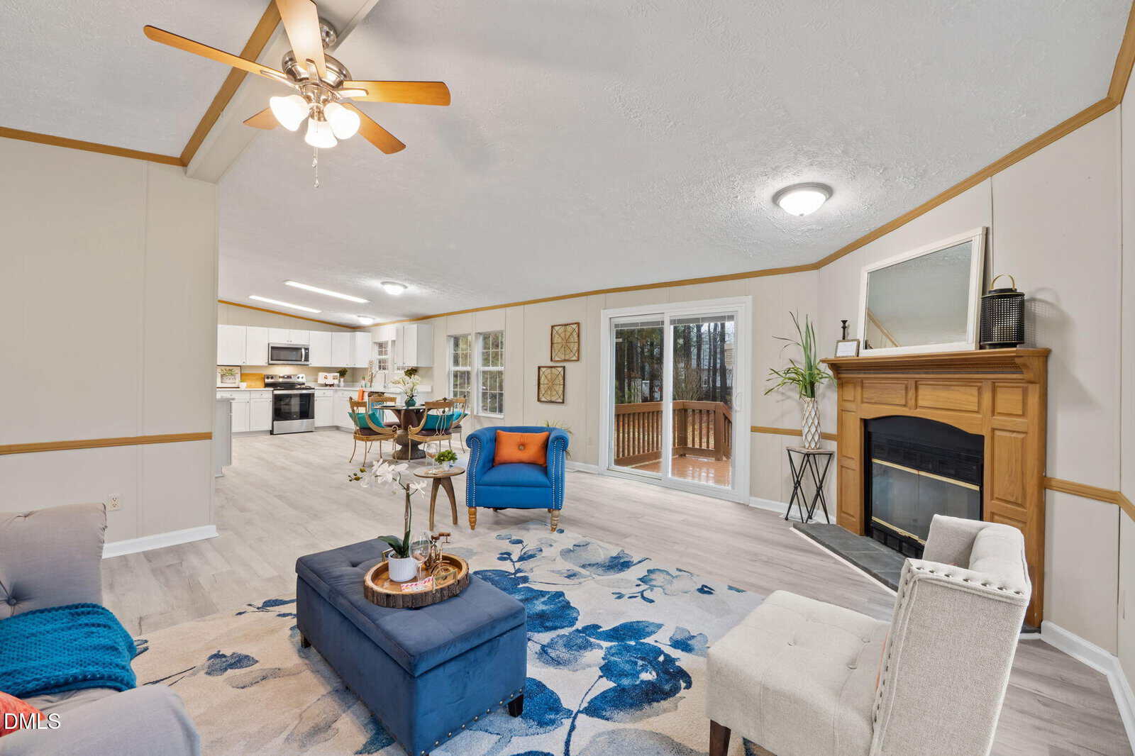 A living room with light walls and wood trim, featuring a fireplace, blue and beige furniture, a patterned rug, and an ottoman. The space opens to a kitchen and dining area, with large windows and a sliding door to a deck.