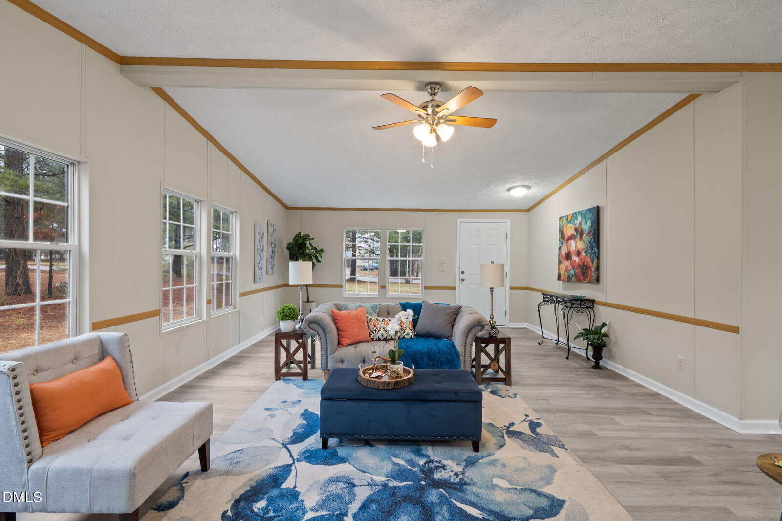 A living room with light-colored walls, wood trim, and wood-look flooring, featuring a gray sofa, a gray armchair with an orange pillow, a blue ottoman, a floral rug, wall art, and a ceiling fan with lights.