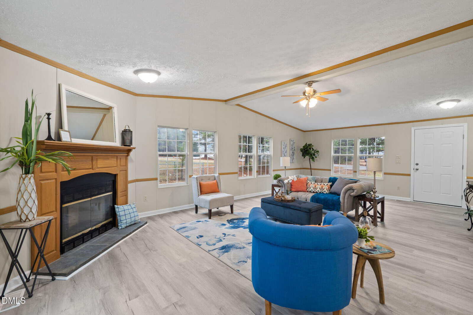 A spacious living room with light wood flooring, a fireplace, blue and orange furniture, a ceiling fan, and several windows letting in natural light. A small table with flowers is beside a blue armchair.