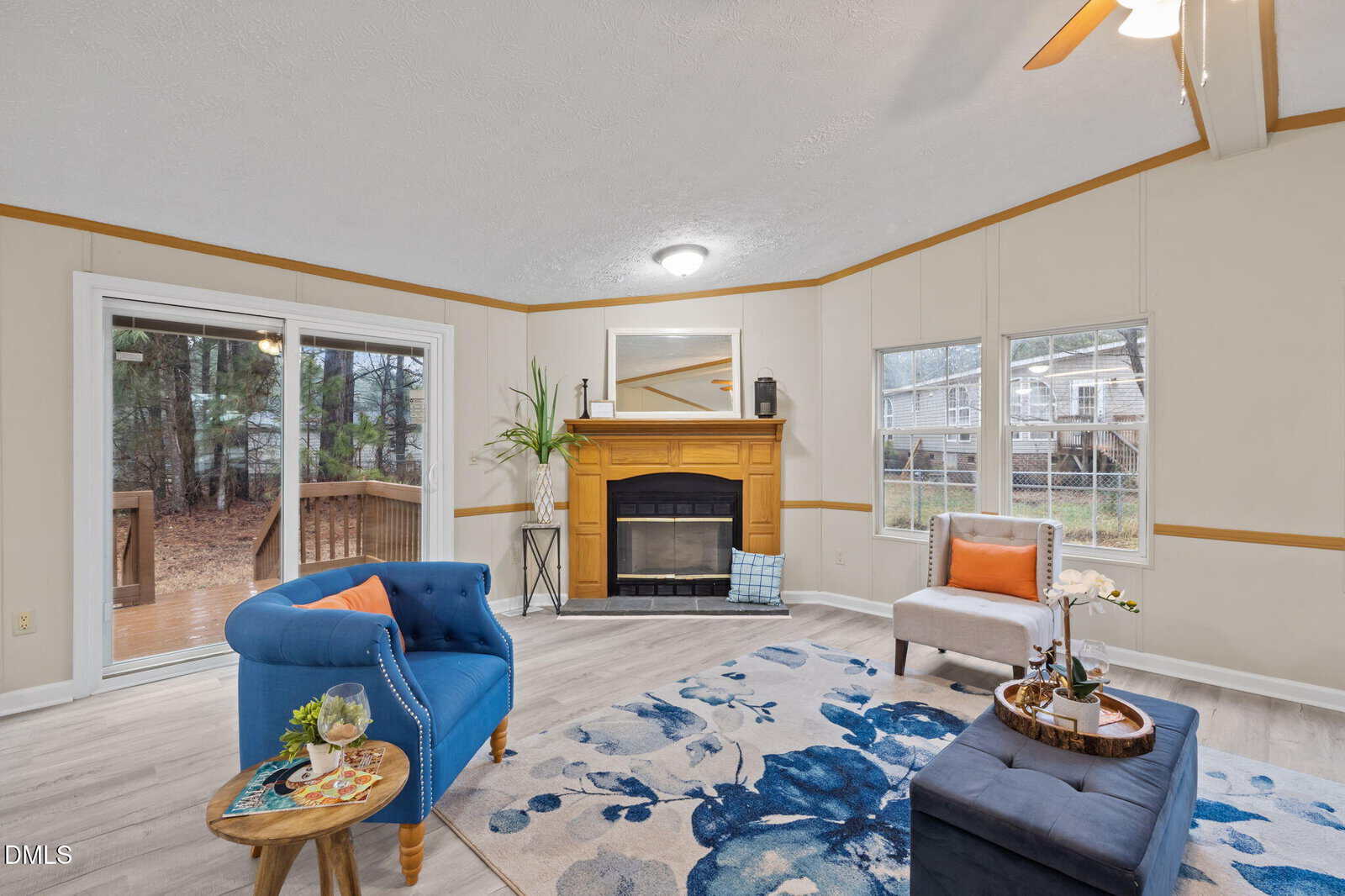 Living room with blue and orange decor, a blue floral rug, armchair, ottoman, and a fireplace with a yellow mantel. Sliding glass door leads to a deck; large windows provide natural light and a view of trees.