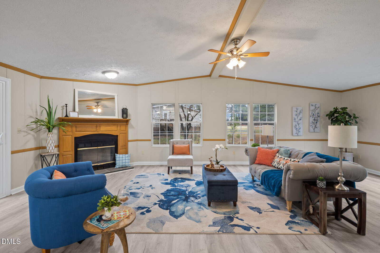 Living room with a blue chair, beige sofa, floral rug, wooden fireplace, ceiling fans, a large window, and wall art. Decor includes plants, cushions, and a small coffee table with books and a drink.