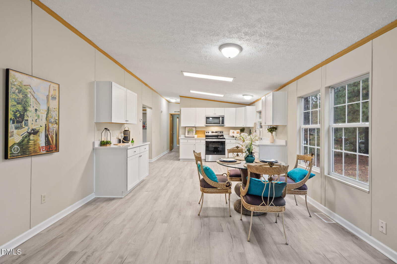 Open kitchen and dining area with light wood flooring, white cabinets, stainless steel appliances, round table with four wicker chairs, large windows on the right, and a framed painting on the left wall.
