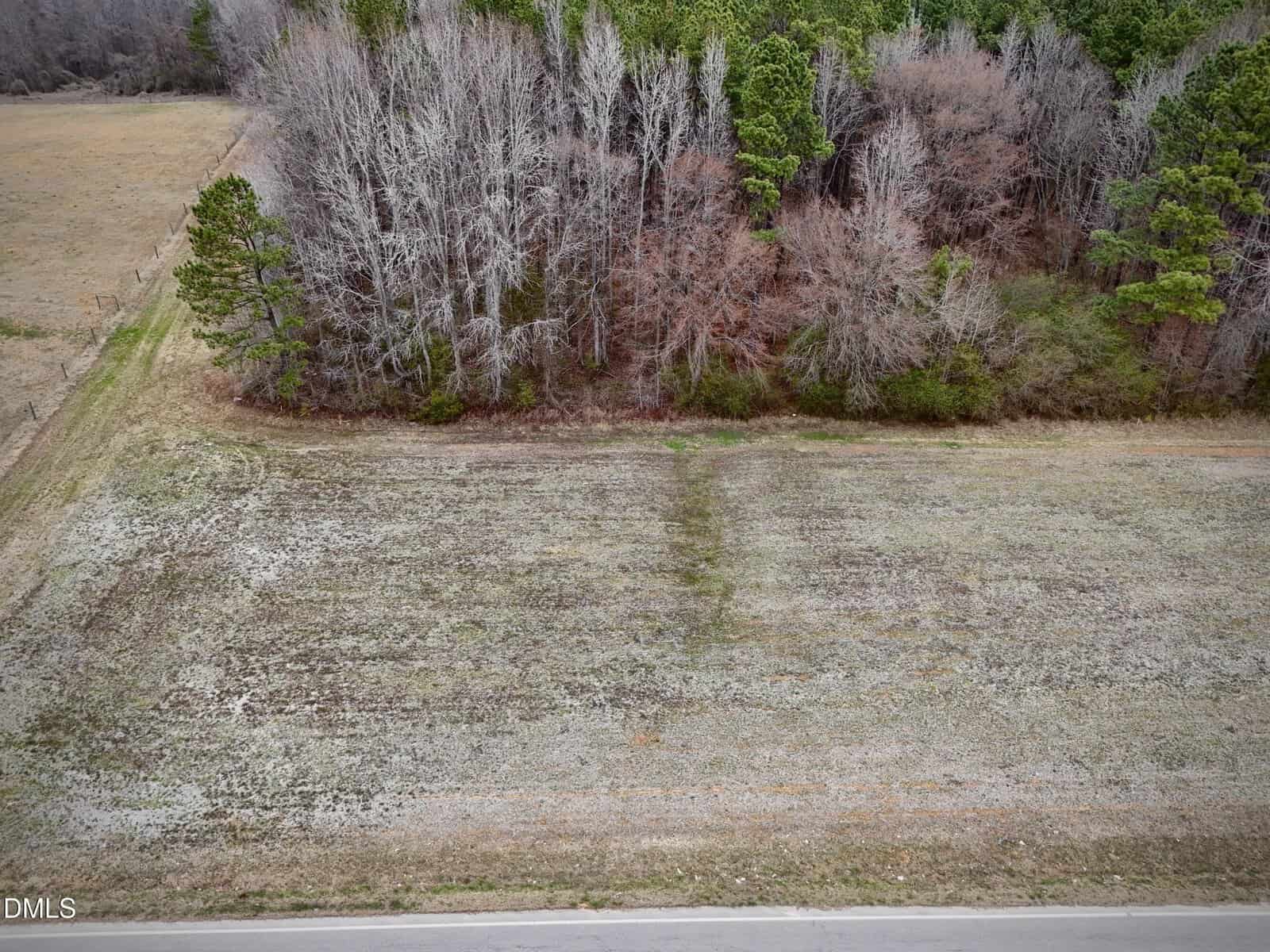 An open, grassy field on Lot 1 W River Road in Franklinton, bordered by a paved road in the foreground and a line of leafless and evergreen trees at the edge of the property in the background.