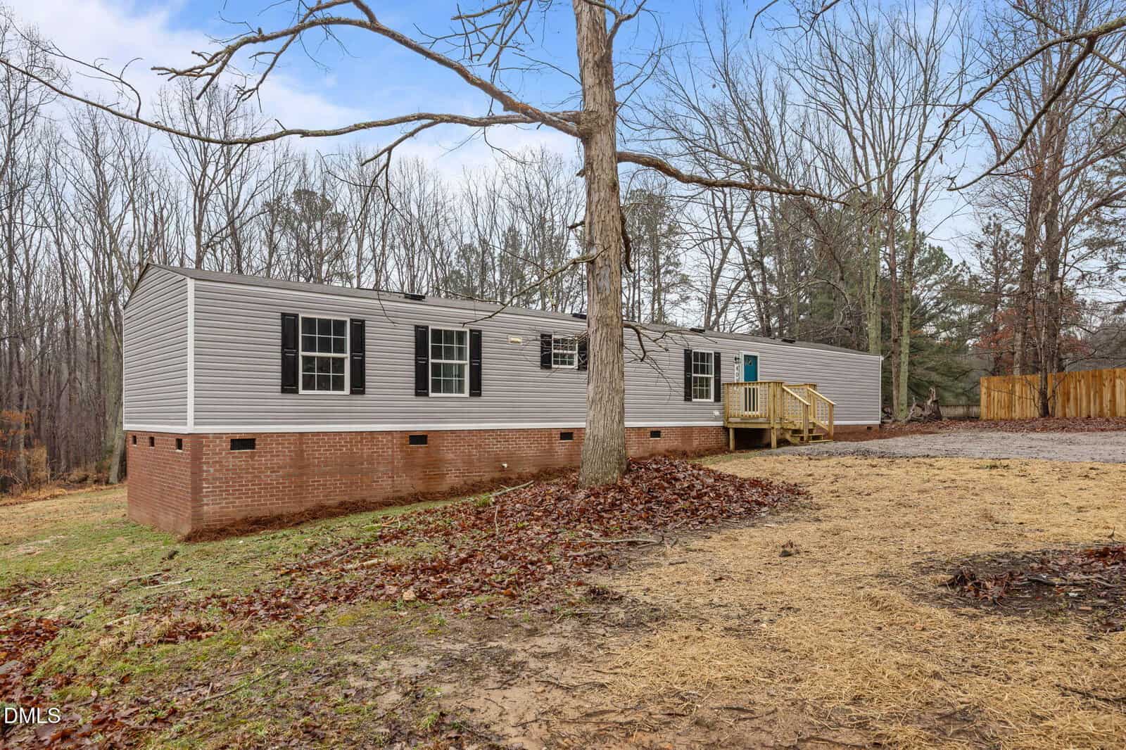 A single-story manufactured home with gray siding and black shutters sits on a brick foundation. There is a wooden ramp leading to the entrance. The yard is partially covered with leaves and bare trees are visible in the background.