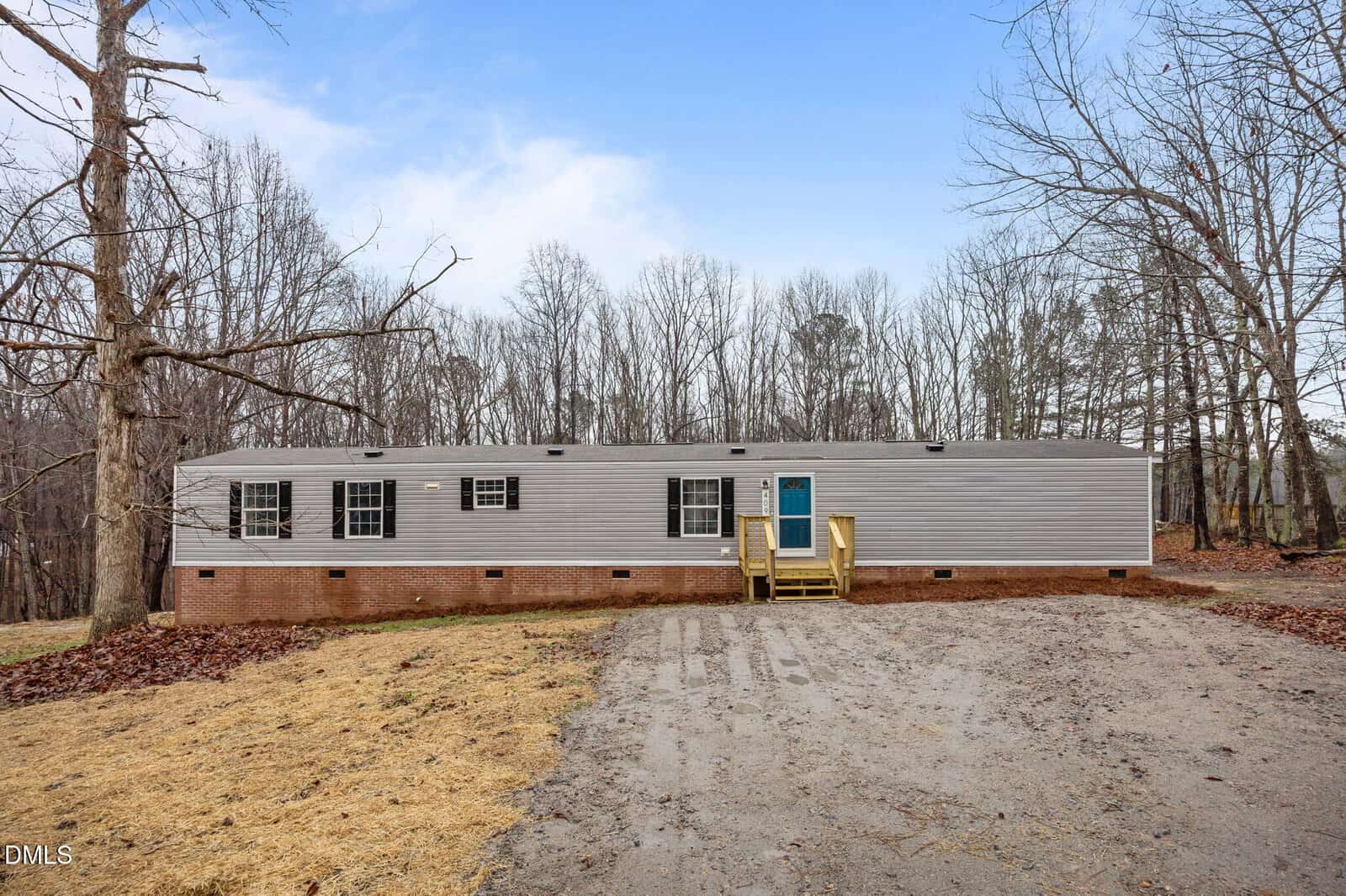 Single-story manufactured home with gray siding, brick foundation, blue front door, wooden steps, and multiple windows. Located at 4099 Oak Circle in Franklinton, this home sits on a cleared lot with a gravel driveway and is surrounded by leafless trees.