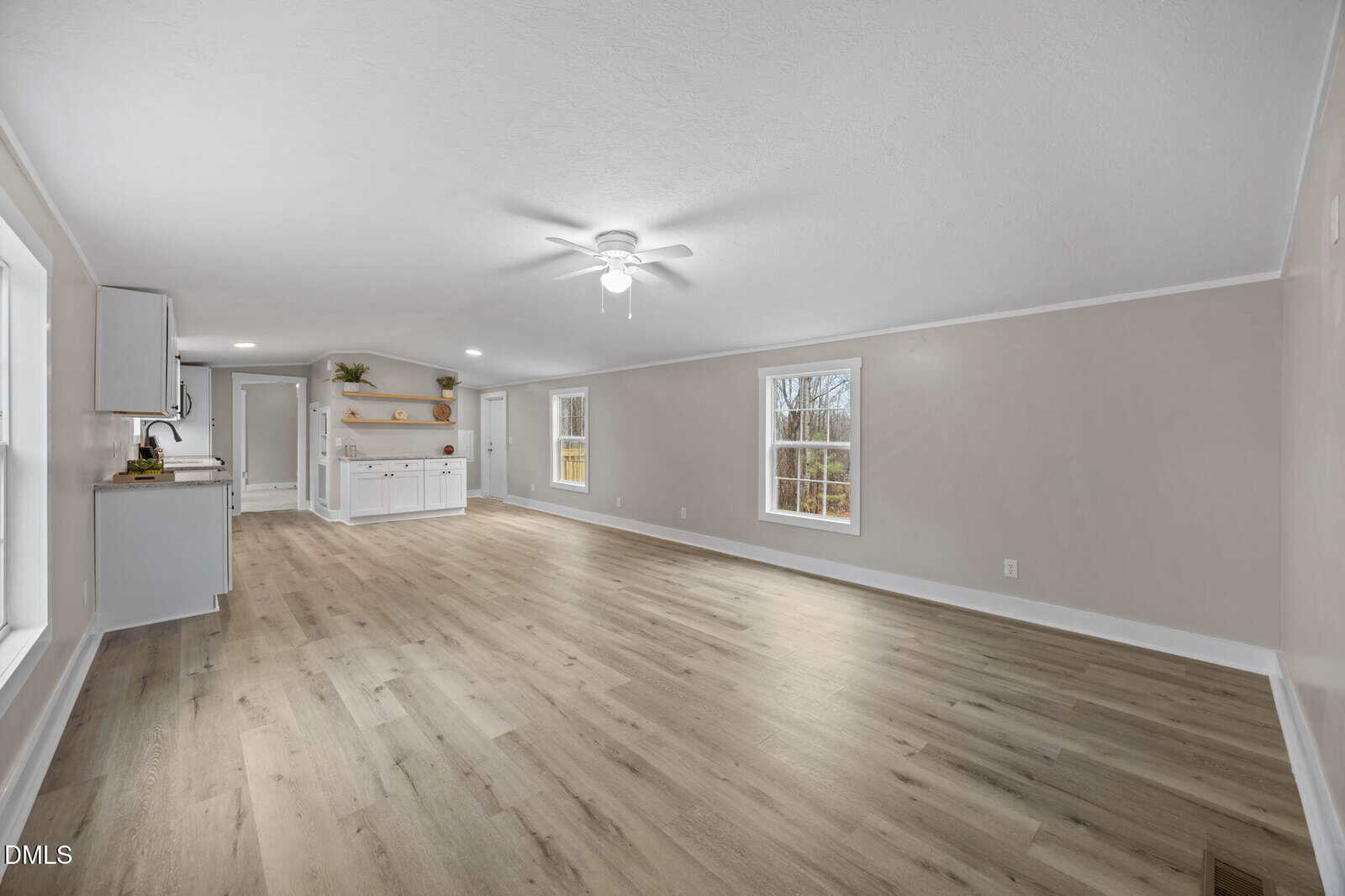 Spacious, empty room with light wood floors, beige walls, white trim, and a ceiling fan. Large windows let in natural light. An open kitchen area with white cabinets and shelves is visible in the background.