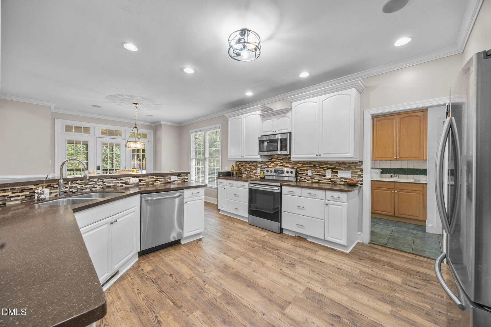 A modern kitchen with white cabinets, stainless steel appliances, a brown countertop, a tile backsplash, and wood flooring. There is a window above the sink and a pendant light over the dining area.