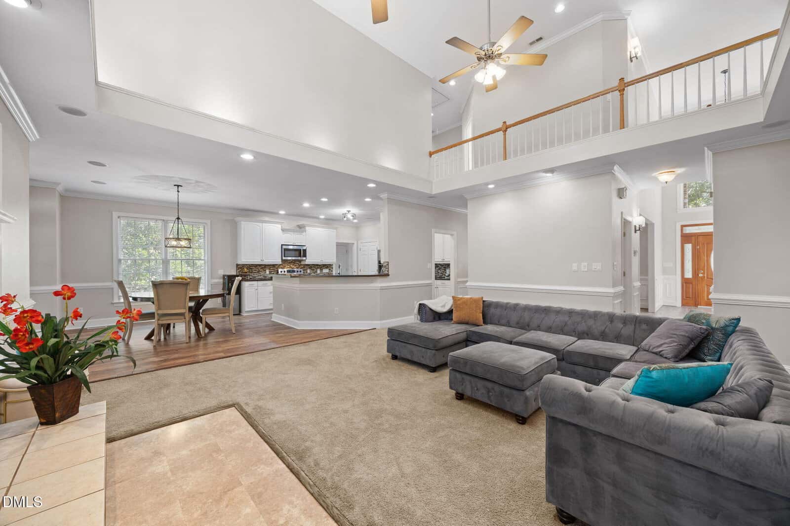 A spacious living room with a gray sectional sofa, beige carpet, and vaulted ceiling with ceiling fans, open to a dining area and white kitchen; large windows provide natural light.