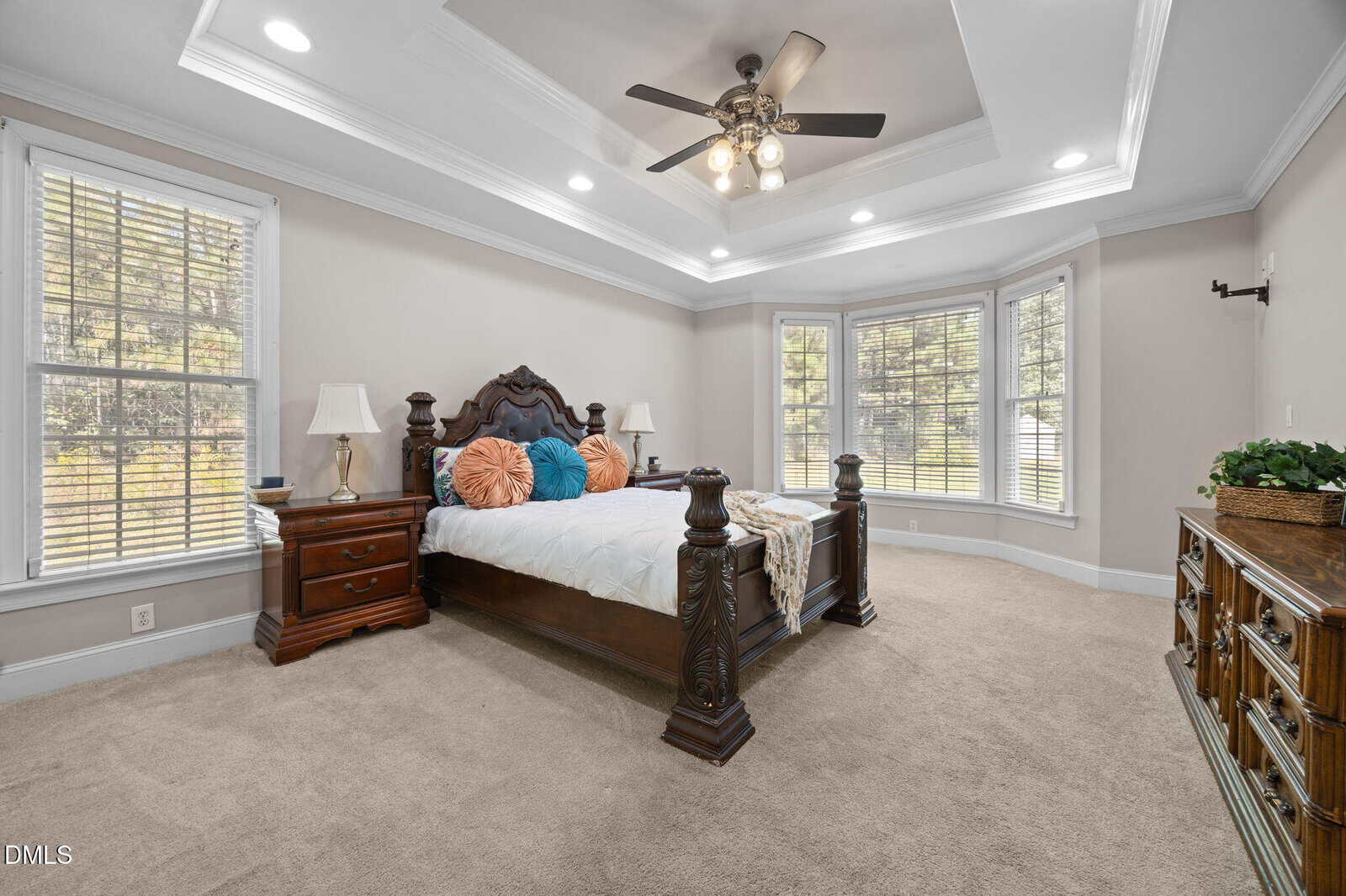 A spacious bedroom with beige carpet, a carved wooden bed with colorful pillows, two nightstands with lamps, a ceiling fan with lights, a dresser with decor, and large windows letting in natural light.