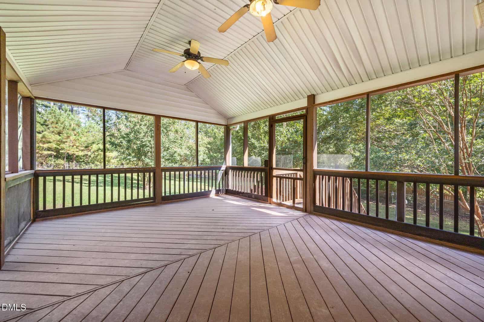 A spacious screened-in porch with a vaulted ceiling, three ceiling fans, and wooden flooring. Large windows offer views of trees and a grassy yard outside.