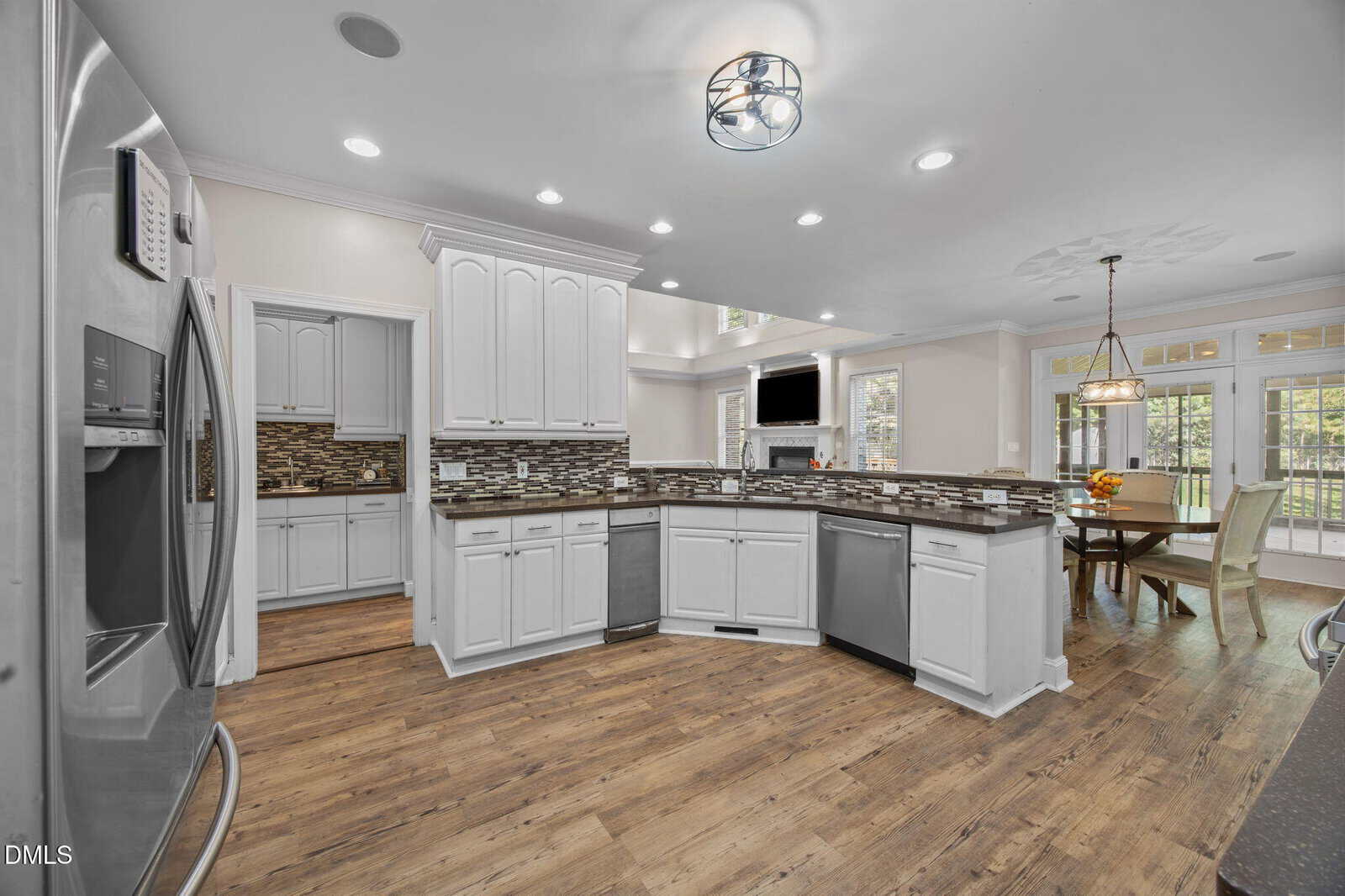 Modern kitchen with white cabinetry, stainless steel appliances, brown backsplash, and wood flooring. An adjacent dining area with a table, chairs, and large windows is visible in the background.
