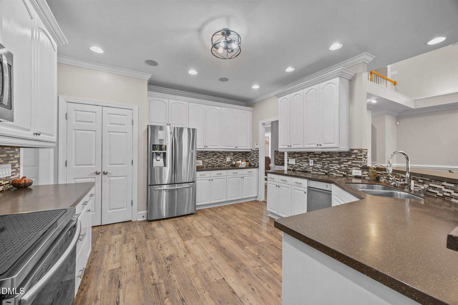 Modern kitchen with white cabinets, stainless steel appliances, brown countertops, mosaic tile backsplash, and wood-style flooring. Recessed ceiling lights and a central ceiling fixture illuminate the space.