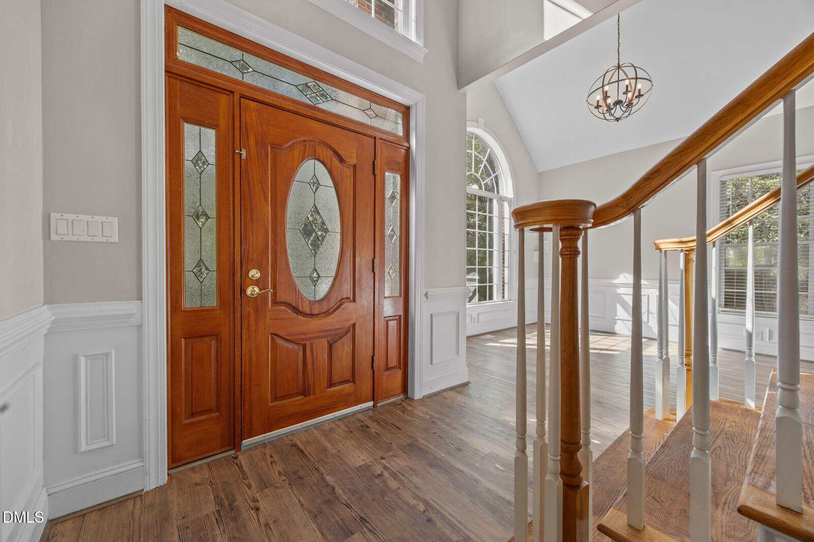 A wooden front door with decorative glass panels is next to a staircase with white spindles and a wooden handrail. Sunlight streams through large windows, illuminating the foyer’s light-colored walls and wood floor.