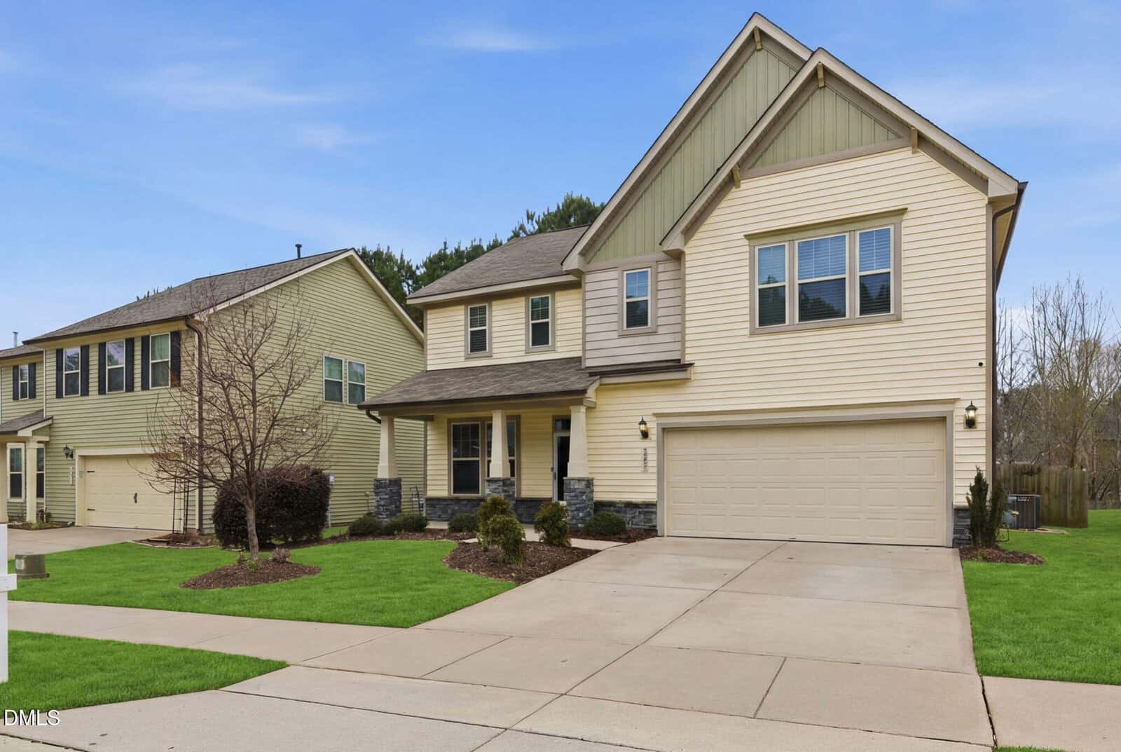 Two-story beige suburban house with a double garage, front porch, and stone accents. Well-kept lawn, small shrubs, and a bare tree in the front yard. Neighboring houses and a clear sky are visible.