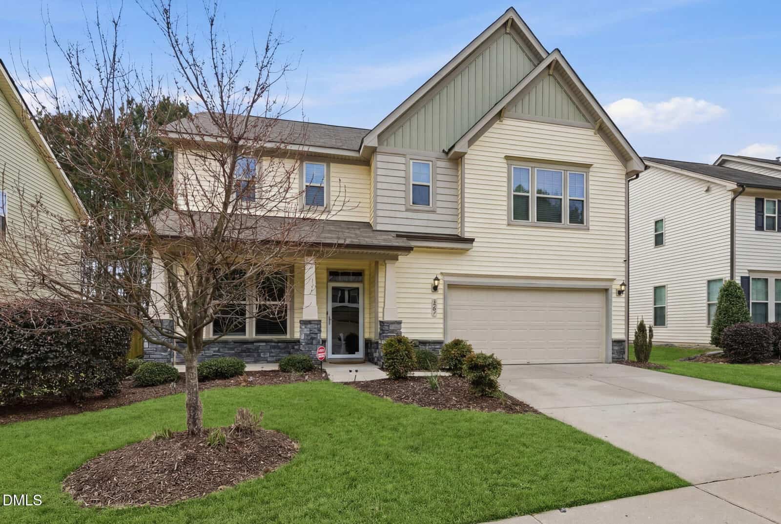 Two-story suburban house with beige siding, green accents, stone trim, and attached two-car garage. Manicured lawn, landscaped shrubs, a bare tree in front yard, and concrete driveway. Blue sky and neighboring homes visible.