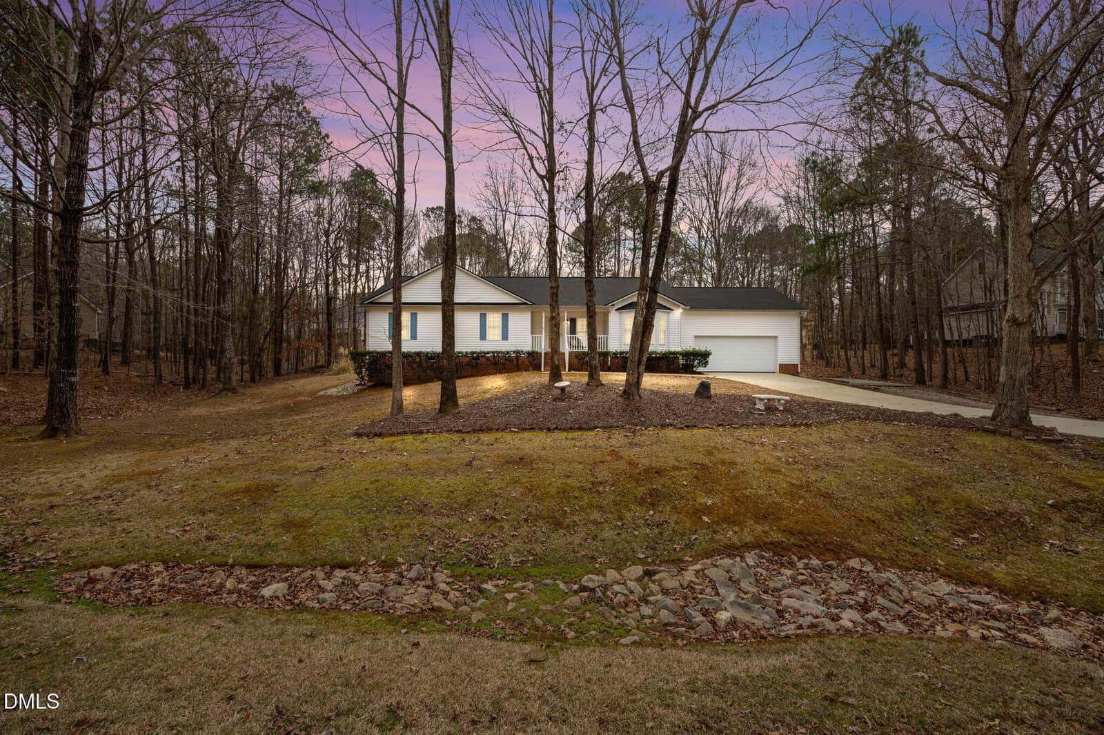 Single-story white house with black shutters and a two-car garage, set among tall, leafless trees at 120 Woodcroft Drive in Youngsville. The front yard is grassy with a stone path and mulched areas, all beneath a purple and pink sky at dusk.