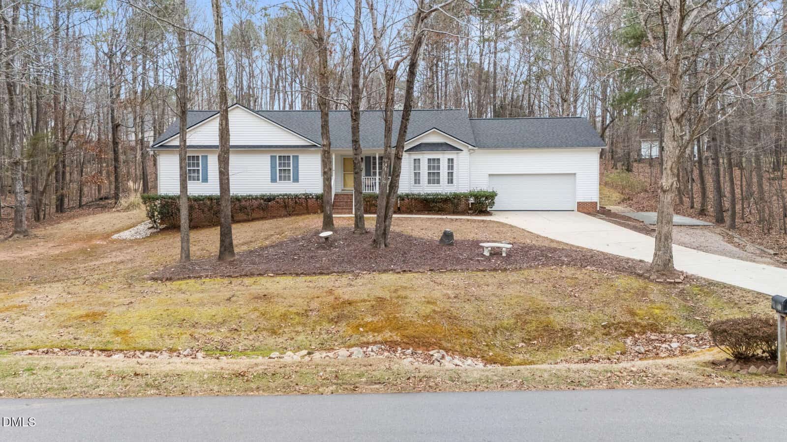 Single-story white house with dark roof, front porch, and attached garage. Several leafless trees in the front yard, paved driveway, and surrounding wooded area. Grass and mulch landscaping visible by the roadside.