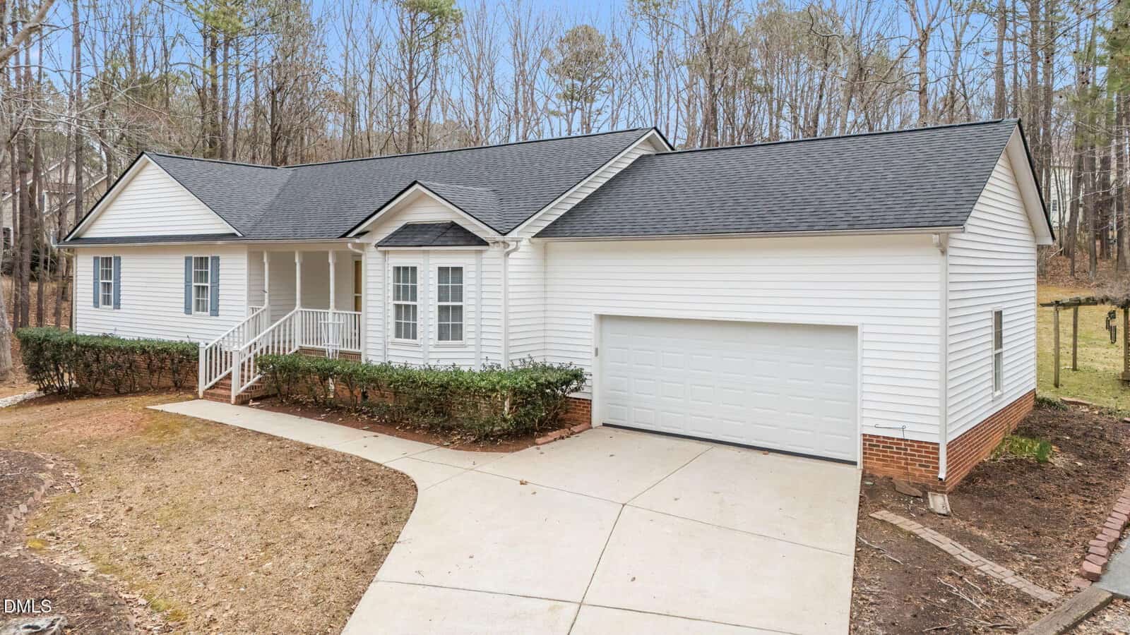 Single-story white house with black roof, attached two-car garage, small front porch with white railing, hedges along the front, concrete driveway, and a wooded area in the background.