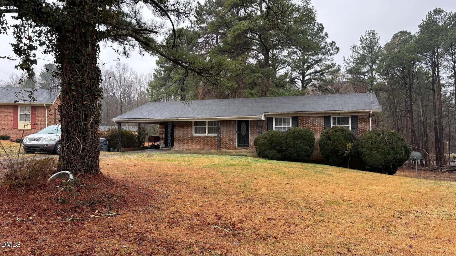 A one-story brick house at 603 Sagefield Way sits on a sloped lawn with brown grass in Henderson. Large shrubs line the front, and pine trees are in the background. A covered carport is attached, with a car parked beside the house beneath an overcast sky.