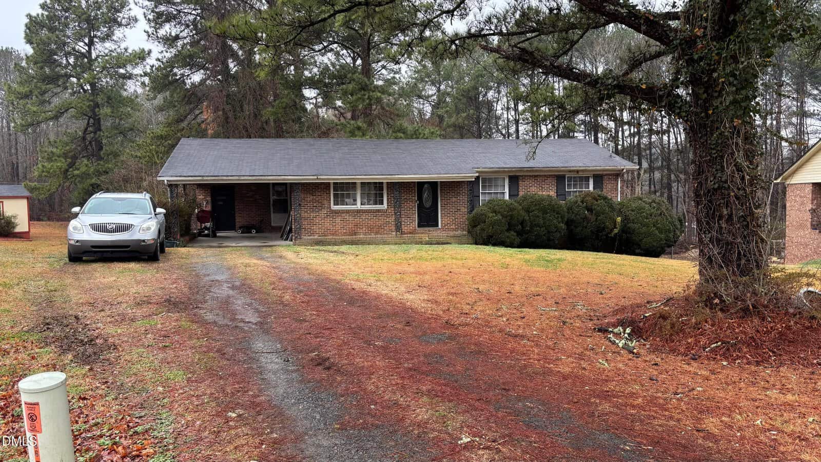 A single-story brick house with a dark roof, two front doors, shrubs by the entrance, and a car parked under a covered carport. The yard has patchy grass and trees in the background. A paved driveway leads to the house.