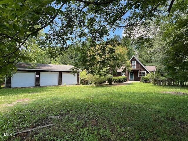 A brown wooden house with a steep roof sits behind a large grassy yard. To the left, there is a detached three-car garage. Trees and greenery surround the property under a partly cloudy sky.