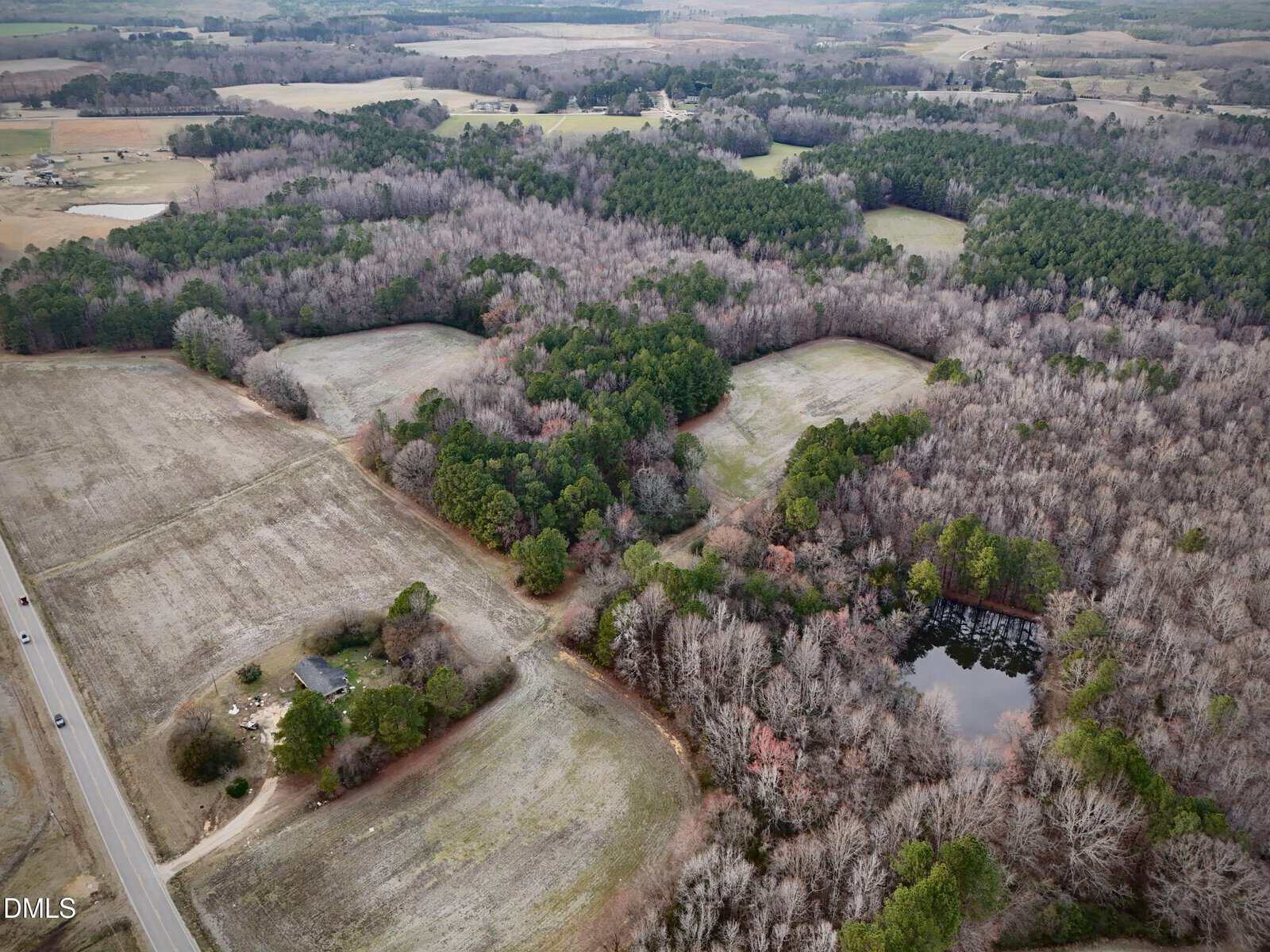 Aerial view of a rural landscape with fields, dense wooded areas, a pond surrounded by trees, scattered houses, and a road running along the left side. The image shows winter foliage with some green pine trees.