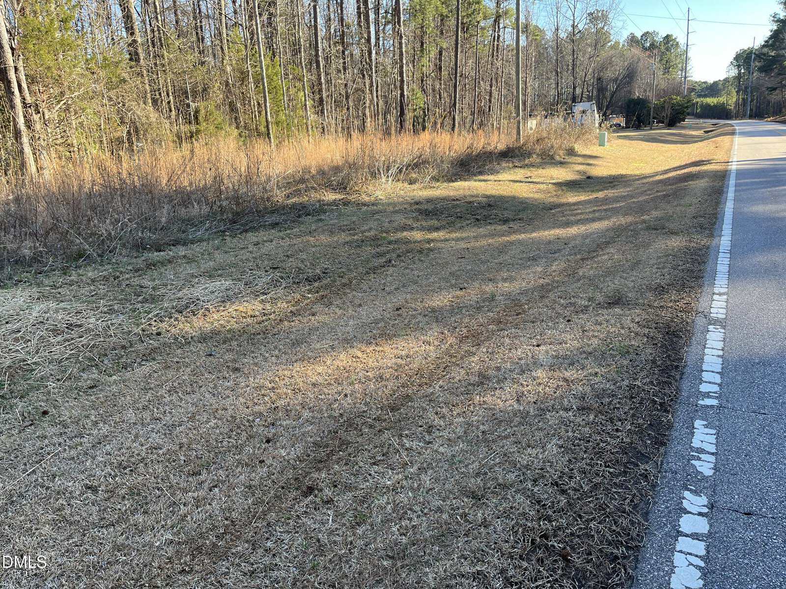 A rural roadside along 00 Nc Hwy 58 near Warrenton features brown grass, tire tracks, trees in the background, and a clear sky. The road curves to the right, with some road signs visible in the distance.