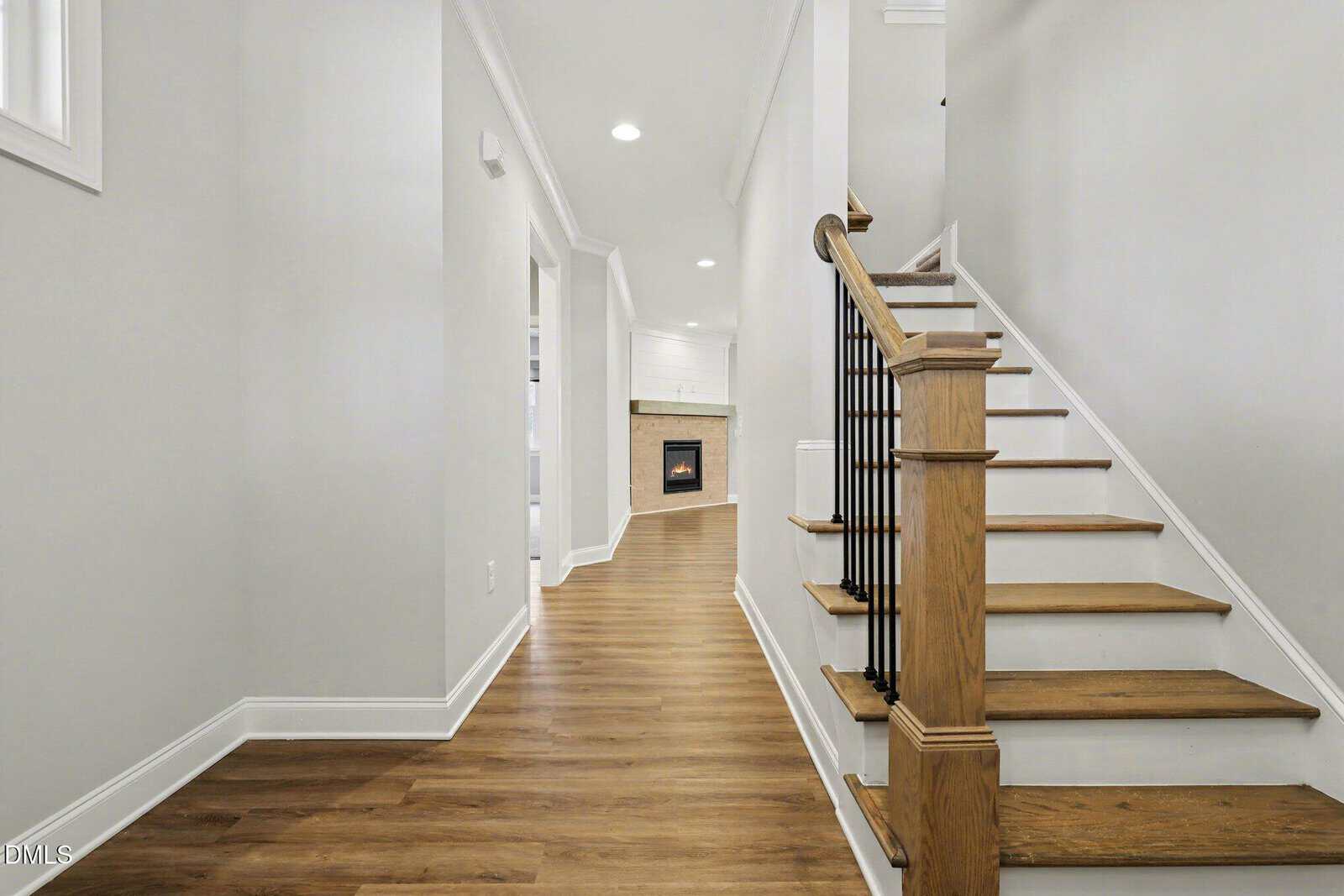 A hallway with light-colored walls and wooden floors leads to a room with a fireplace. Wooden stairs with a black railing are on the right, and recessed ceiling lights brighten the space.