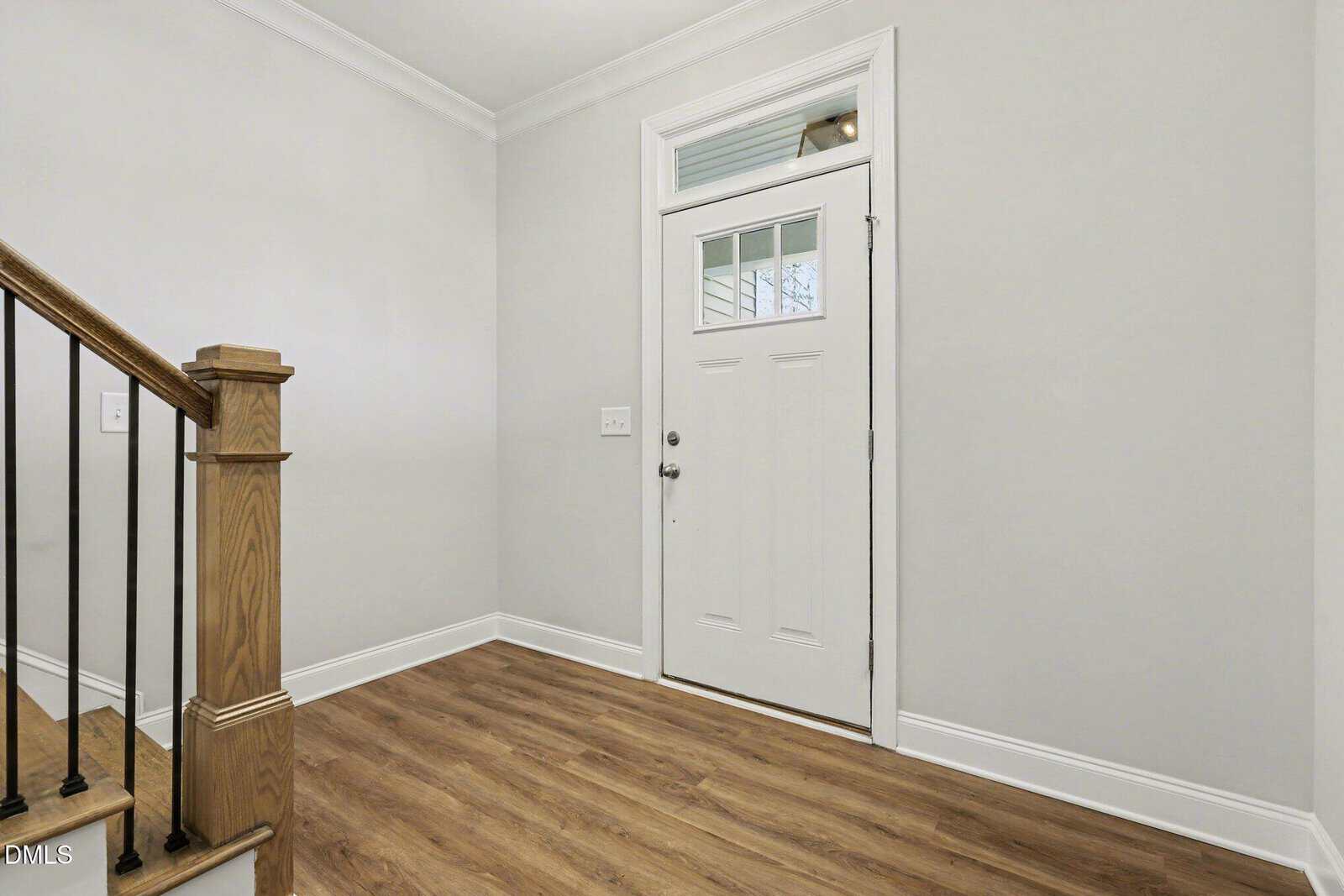 A simple, clean entryway with light gray walls, white trim, wood-look flooring, and a white front door with a transom window above. A staircase with a wooden handrail and black spindles is visible on the left.