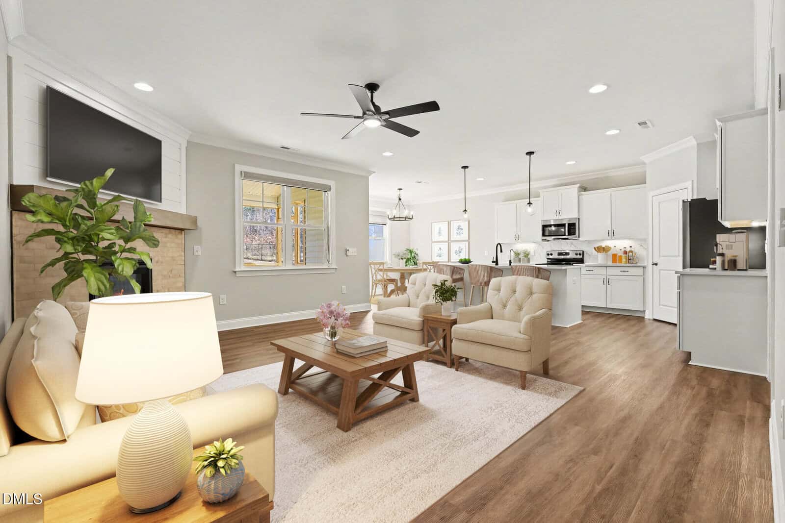A bright, open-concept living room and kitchen with light wood floors, neutral-toned furniture, a ceiling fan, built-in fireplace, indoor plants, and modern stainless steel appliances.